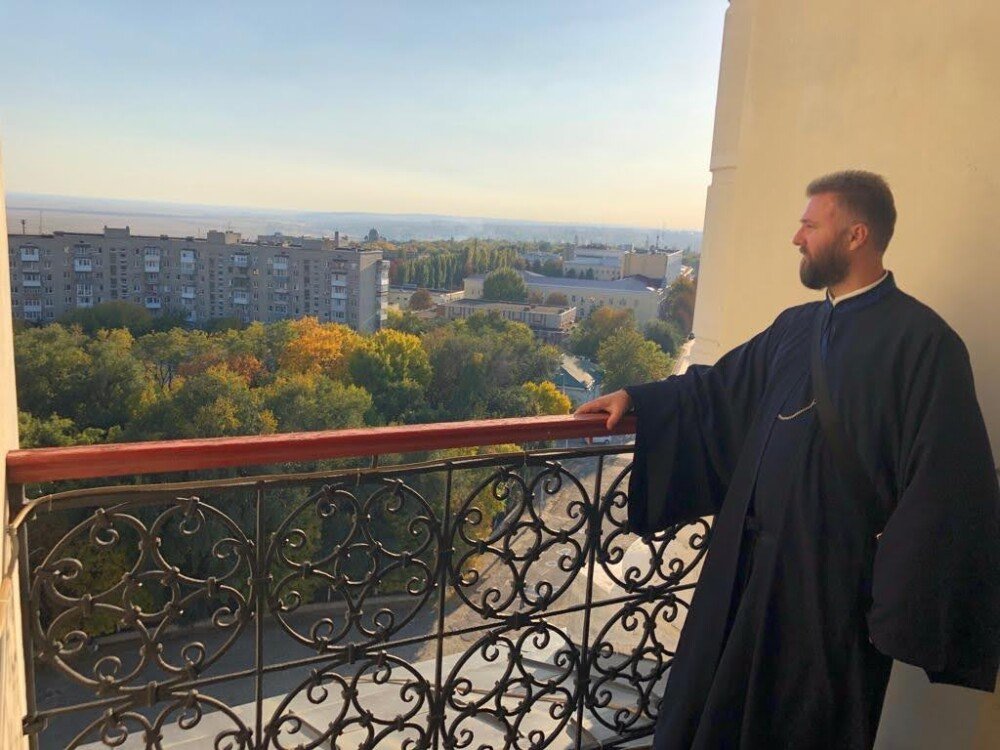 Отац Предраг Поповић | Brojanice Milojević Човек у тамној хаљини, са бројаницом као духовним накитом, стоји на балкону са украшеном гвозденом оградом, посматрајући градски пејзаж дрвећа и зграда под ведрим небом.