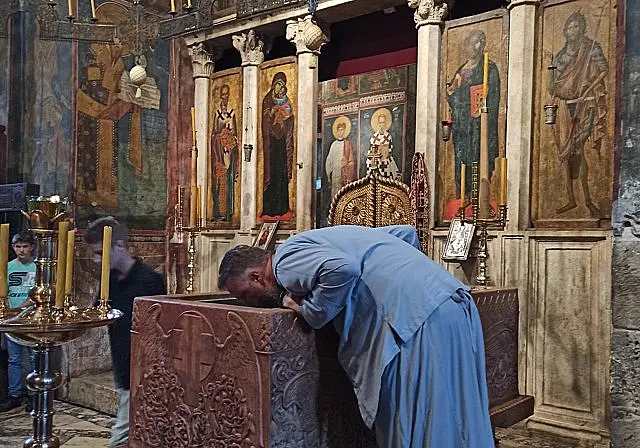 Човек у верској одећи са бројаницом нагиње се и љуби богато украшени камени олтар у цркви, док други посматрају. Иза њега су осликане иконе и златни свећњаци.