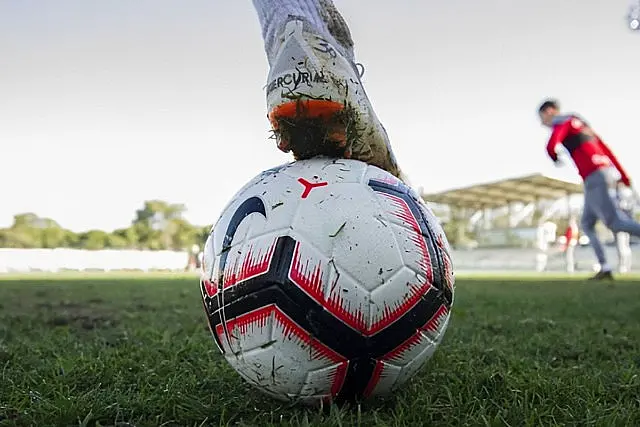 Крупни план стопала фудбалера у белој копачки и сребрној бројаници, како лежи на фудбалској лопти на травнатом терену, са другим играчем и стадионом видљивим у замагљеној позадини.