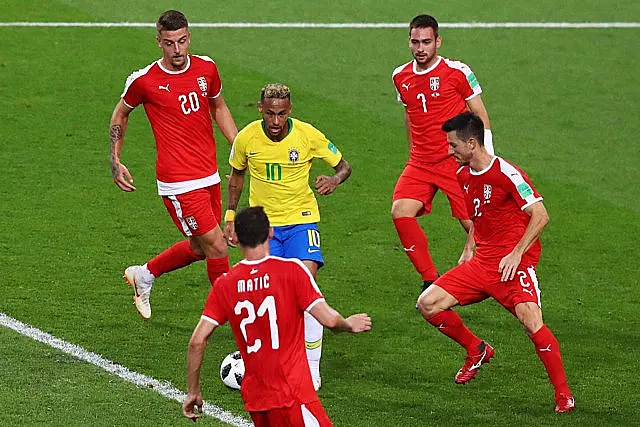 Бразилски фудбалер у жутом дресу дрибла лопту, покрети су му готово духовити, док је окружен четворицом српских играча у црвеним униформама током утакмице на зеленом терену.